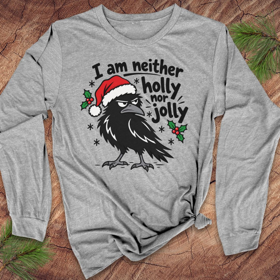 Athletic heather gray long-sleeve t-shirt with a grumpy raven in a Santa hat surrounded by holly and snowflakes and the words I am neither holly nor jolly, laid flat on a rustic wood background with pine branches.