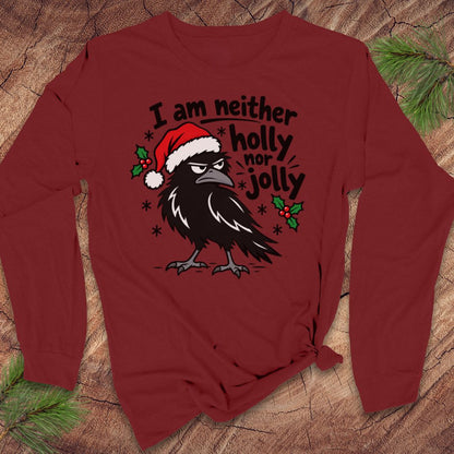 Cardinal red long-sleeve t-shirt with a grumpy raven in a Santa hat surrounded by holly and snowflakes and the words I am neither holly nor jolly, laid flat on a rustic wood background with pine branches.