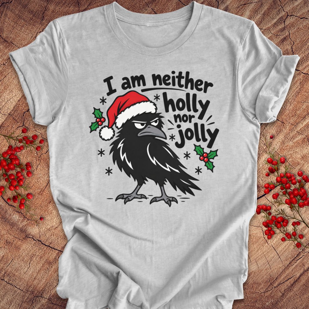 Athletic heather gray t-shirt with a grumpy raven in a Santa hat surrounded by holly and snowflakes and the words I am neither holly nor jolly, laid flat on a rustic wood background with red winter berries.
