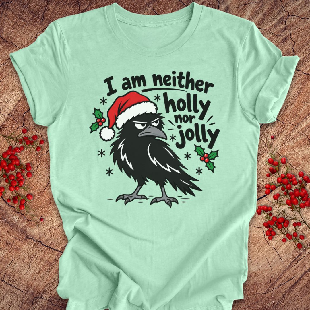 Heather mint green t-shirt with a grumpy raven in a Santa hat surrounded by holly and snowflakes and the words I am neither holly nor jolly, laid flat on a rustic wood background with red winter berries.