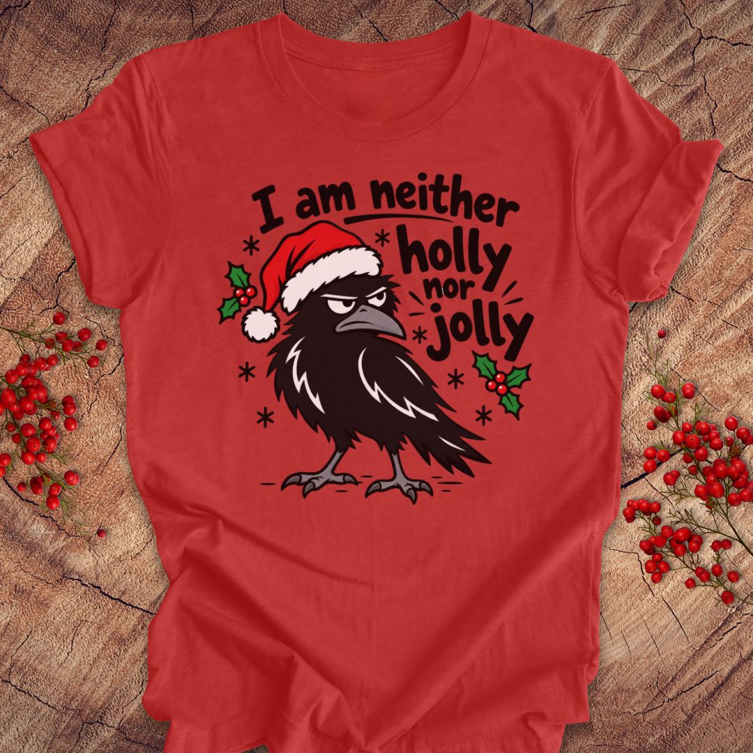 Heather red t-shirt with a grumpy raven in a Santa hat surrounded by holly and snowflakes and the words I am neither holly nor jolly, laid flat on a rustic wood background with red winter berries.