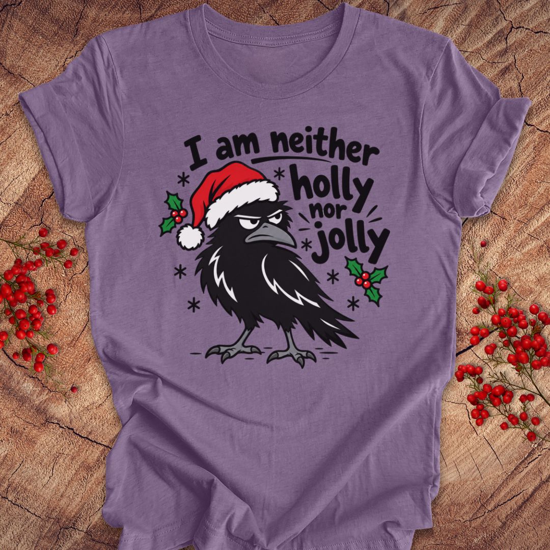 Heather purple t-shirt with a grumpy raven in a Santa hat surrounded by holly and snowflakes and the words I am neither holly nor jolly, laid flat on a rustic wood background with red winter berries.