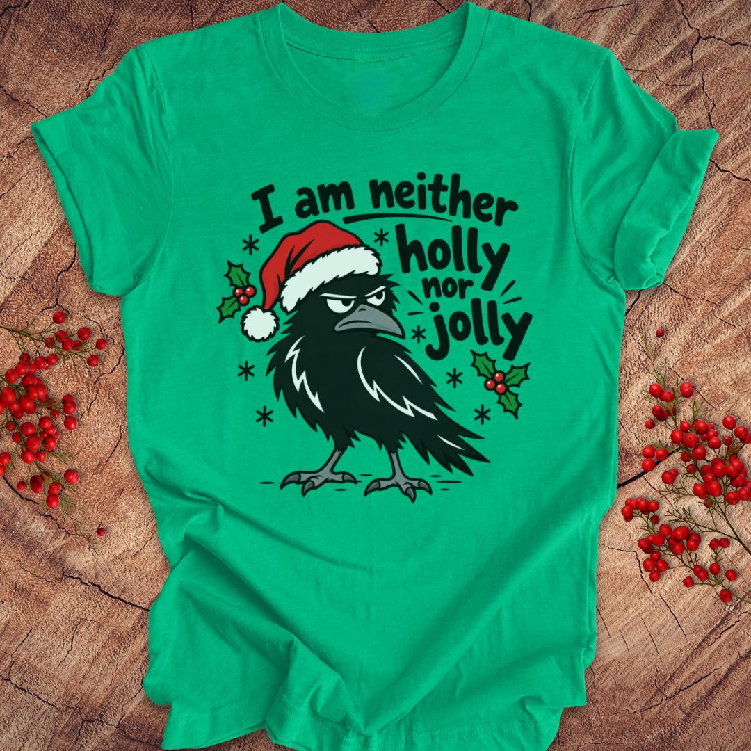 Heather kelly green t-shirt with a grumpy raven in a Santa hat surrounded by holly and snowflakes and the words I am neither holly nor jolly, laid flat on a rustic wood background with red winter berries.