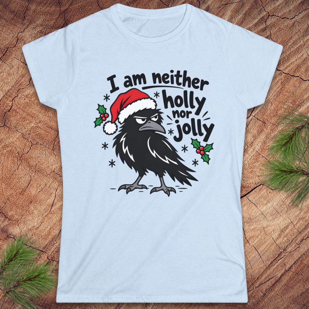 Light blue women's t-shirt with a grumpy raven in a Santa hat surrounded by holly and snowflakes and the words I am neither holly nor jolly, laid flat on a rustic wood background with pine branches.
