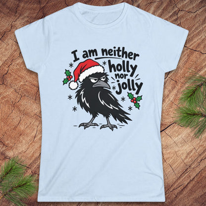 Light blue women's t-shirt with a grumpy raven in a Santa hat surrounded by holly and snowflakes and the words I am neither holly nor jolly, laid flat on a rustic wood background with pine branches.