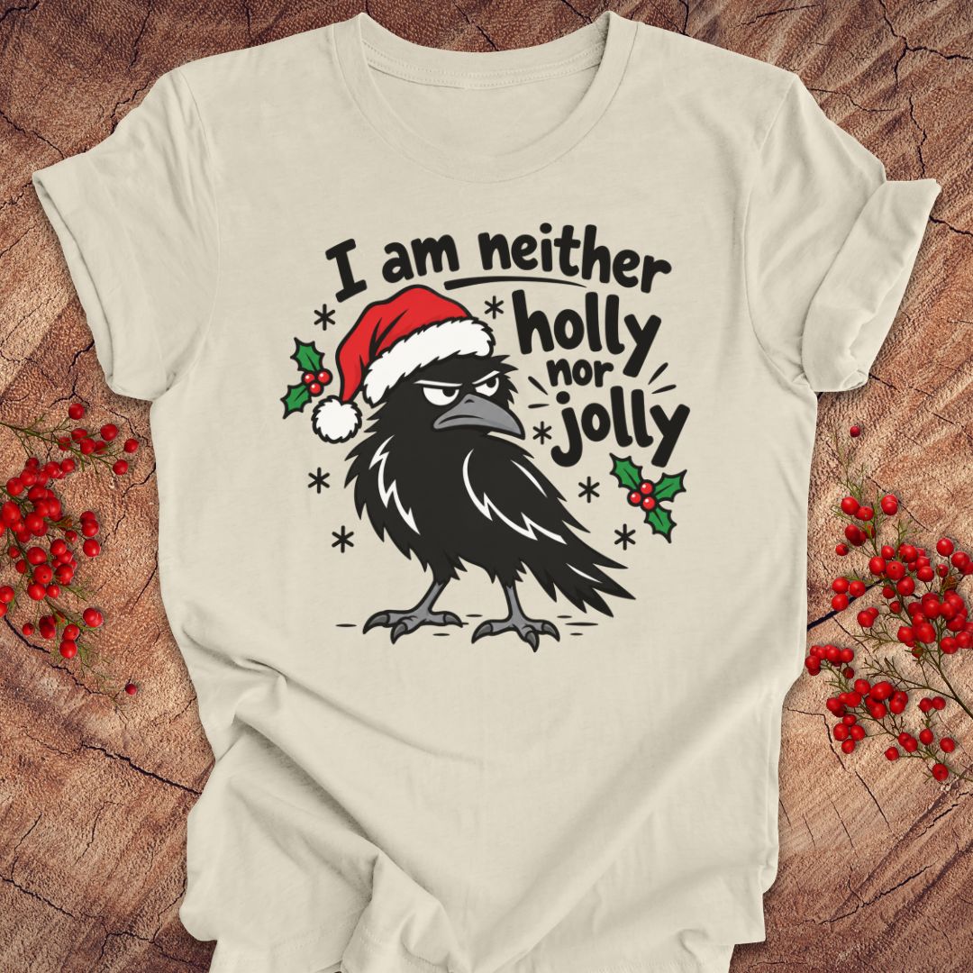 Natural cream t-shirt with a grumpy raven in a Santa hat surrounded by holly and snowflakes and the words I am neither holly nor jolly, laid flat on a rustic wood background with red winter berries.