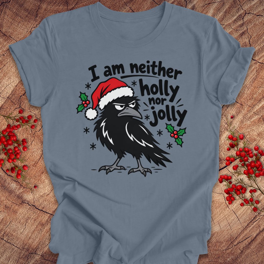 Steel blue t-shirt with a grumpy raven in a Santa hat surrounded by holly and snowflakes and the words I am neither holly nor jolly, laid flat on a rustic wood background with red winter berries.