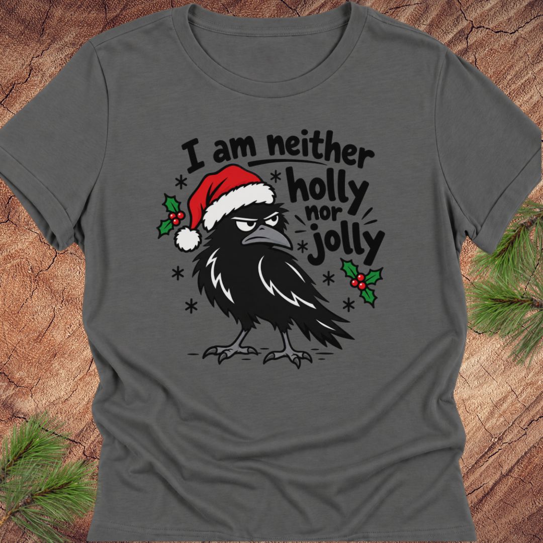 Asphalt gray women's relaxed t-shirt with a grumpy raven in a Santa hat surrounded by holly and snowflakes and the words I am neither holly nor jolly, laid flat on a rustic wood background with pine branches.
