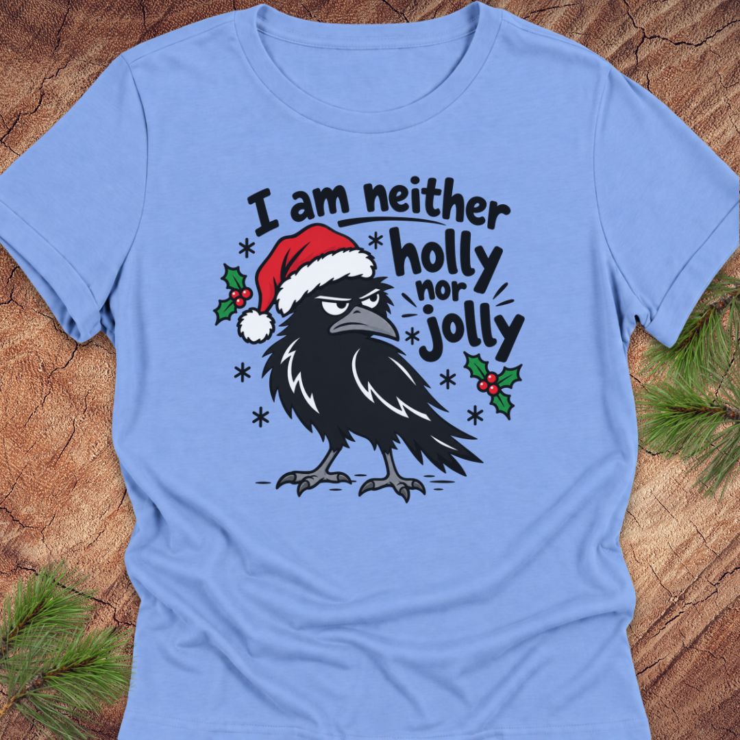 Carolina blue women's relaxed t-shirt with a grumpy raven in a Santa hat surrounded by holly and snowflakes and the words I am neither holly nor jolly, laid flat on a rustic wood background with pine branches.