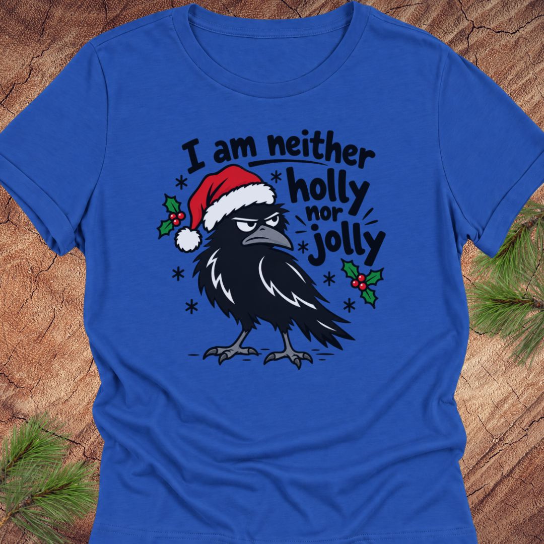 True royal blue women's relaxed t-shirt with a grumpy raven in a Santa hat surrounded by holly and snowflakes and the words I am neither holly nor jolly, laid flat on a rustic wood background with pine branches.