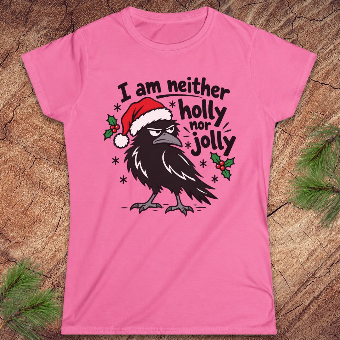 Azalea pink women's t-shirt with a grumpy raven in a Santa hat surrounded by holly and snowflakes and the words I am neither holly nor jolly, laid flat on a rustic wood background with pine branches.