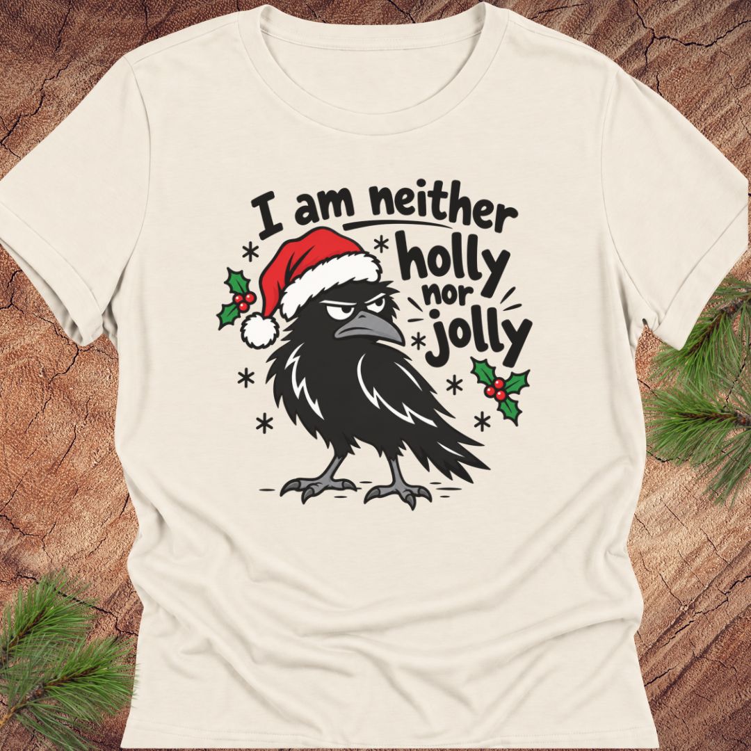 Natural cream women's relaxed t-shirt with a grumpy raven in a Santa hat surrounded by holly and snowflakes and the words I am neither holly nor jolly, laid flat on a rustic wood background with pine branches.