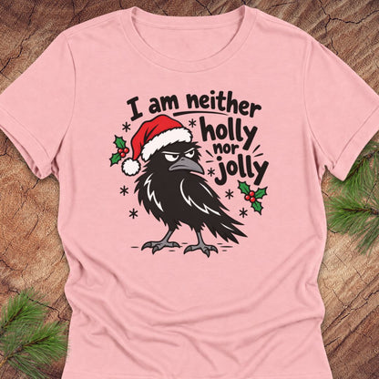 Pink women's relaxed t-shirt with a grumpy raven in a Santa hat surrounded by holly and snowflakes and the words I am neither holly nor jolly, laid flat on a rustic wood background with pine branches.