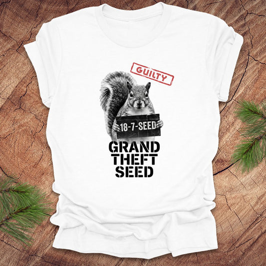 White t-shirt with a squirrel holding a seed packet graphic and text, 'guilty. grand theft seed'on a wooden surface.