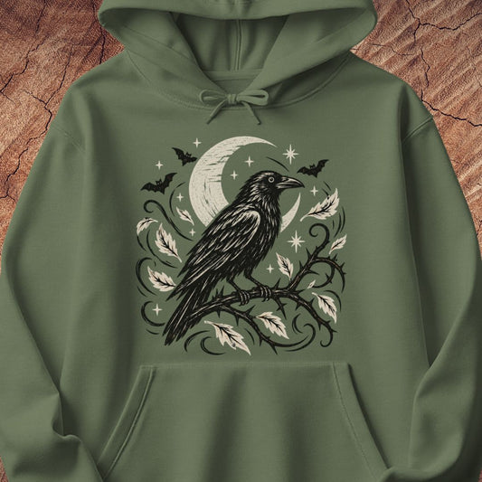 An olive green pullover hoodie featuring a detailed black and white graphic of a raven perched on a branch with leaves, a crescent moon, bats, and stars, displayed against a textured brown wood background.