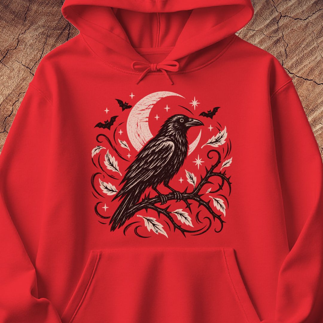 A bright red pullover hoodie laid flat on a rustic wood background, featuring a large front graphic of a black raven perched on a branch with pale pink leaves, a crescent moon, bats, and small stars.