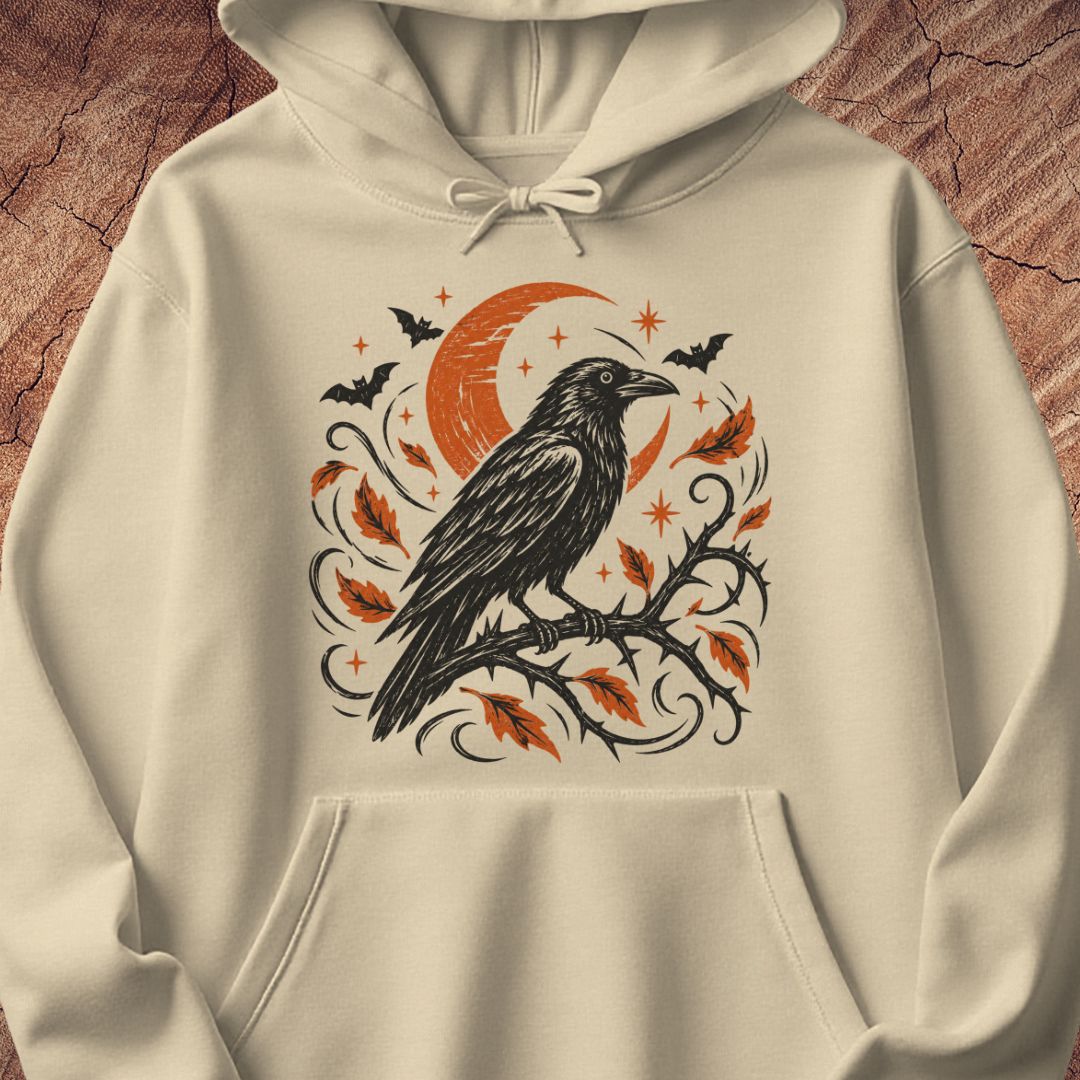 An off-white hooded sweatshirt featuring a detailed graphic print of a black raven perched on a branch with orange leaves, a crescent moon, and bats, displayed against a textured brown wood background.