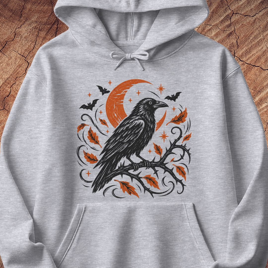 A light heather gray hoodie featuring a Halloween-themed graphic print of a black raven on a branch with orange leaves, a crescent moon, bats, and stars, displayed on a rustic wood background.