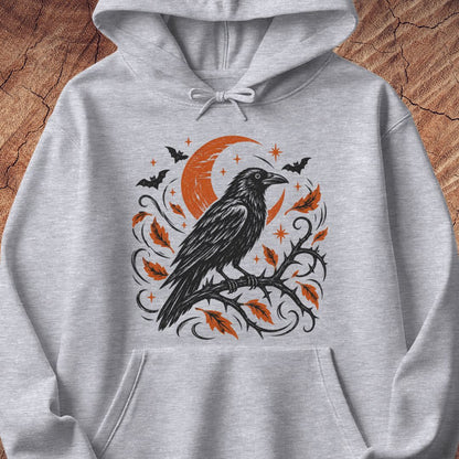 A light heather gray hoodie featuring a Halloween-themed graphic print of a black raven on a branch with orange leaves, a crescent moon, bats, and stars, displayed on a rustic wood background.