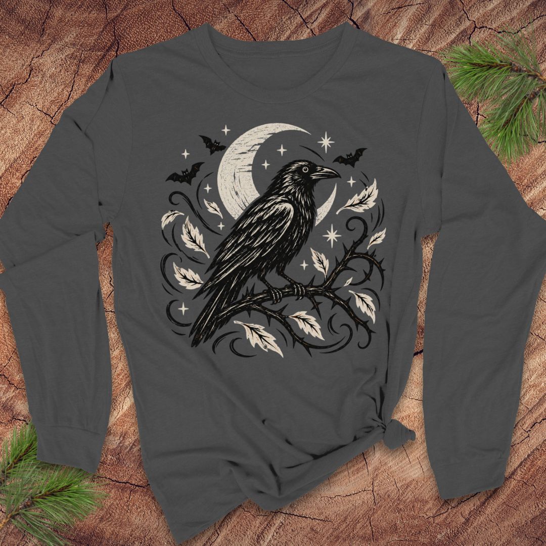 A dark gray long-sleeve t-shirt with a black and white graphic of a raven perched on a branch under a crescent moon and stars, laid flat on a rustic wood background with pine branches in the corners.