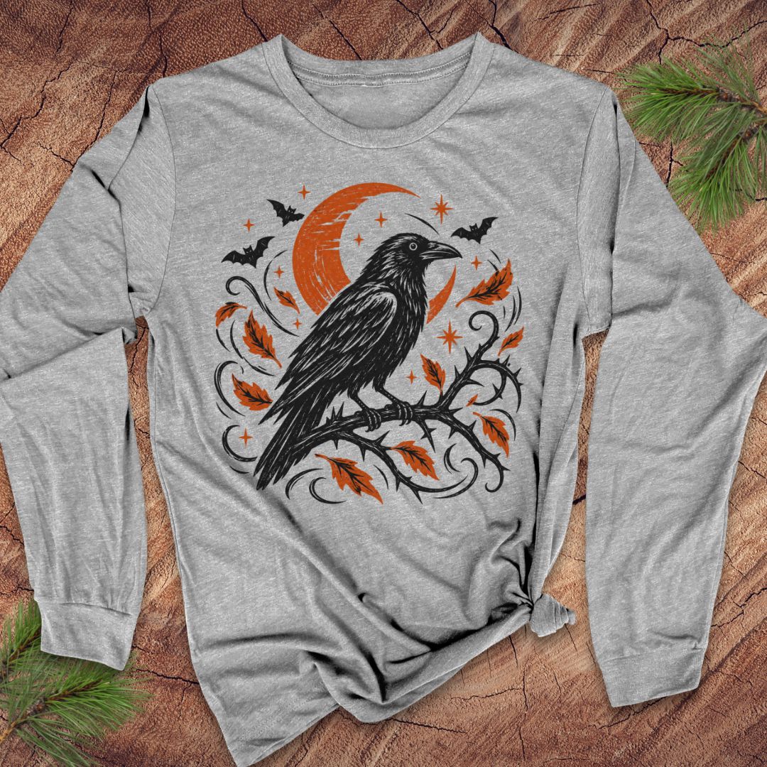 A gray long-sleeve t-shirt with a black and white graphic of a raven perched on a branch under a crescent moon and stars, laid flat on a rustic wood background with pine branches in the corners.