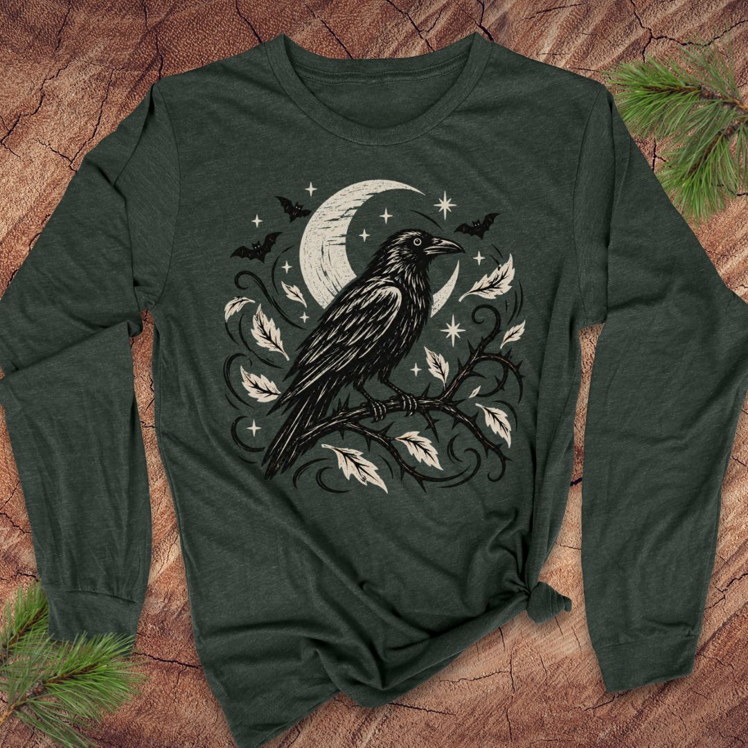 A dark forest green long-sleeve t-shirt with a black and white graphic of a raven perched on a branch under a crescent moon and stars, laid flat on a rustic wood background with pine branches in the corners.
