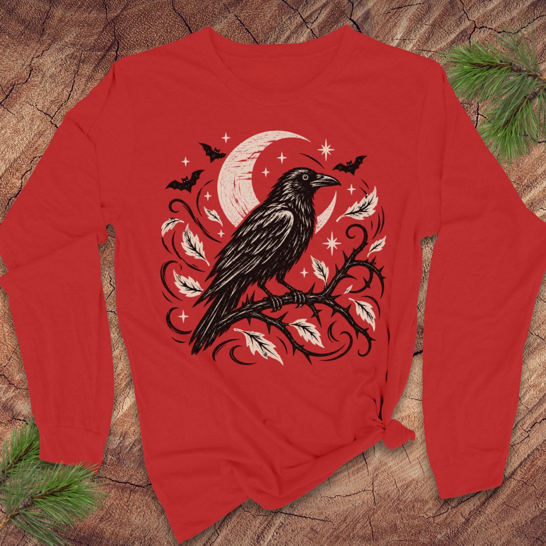 A red long-sleeve t-shirt with a black and white graphic of a raven perched on a branch under a crescent moon and stars, laid flat on a rustic wood background with pine branches in the corners.