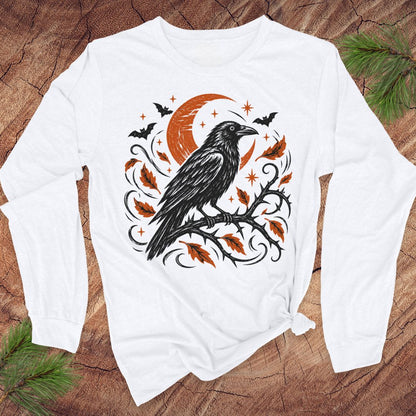 A white long-sleeve t-shirt with a black and white graphic of a raven perched on a branch under a crescent moon and stars, laid flat on a rustic wood background with pine branches in the corners.