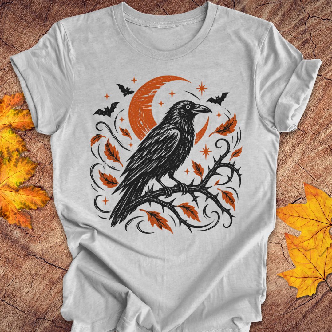 A light gray short-sleeve t-shirt with a Halloween-themed graphic print featuring a black raven on a branch with orange leaves, a crescent moon, bats, and stars. The shirt is displayed flat on a rustic wood background with autumn leaves.