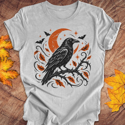 A light gray short-sleeve t-shirt with a Halloween-themed graphic print featuring a black raven on a branch with orange leaves, a crescent moon, bats, and stars. The shirt is displayed flat on a rustic wood background with autumn leaves.