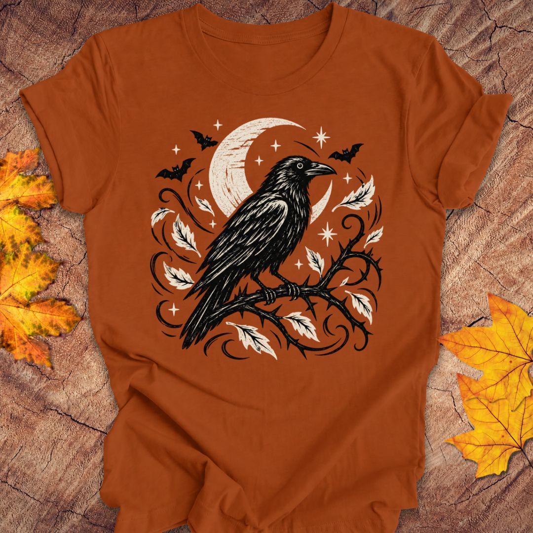 A burn orange short-sleeve t-shirt with a Halloween graphic of a black raven on a branch with orange leaves and a crescent moon, laid flat on a rustic wood background with autumn leaves.