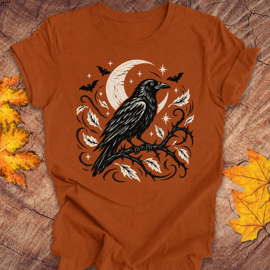 A burn orange short-sleeve t-shirt with a Halloween graphic of a black raven on a branch with orange leaves and a crescent moon, laid flat on a rustic wood background with autumn leaves.