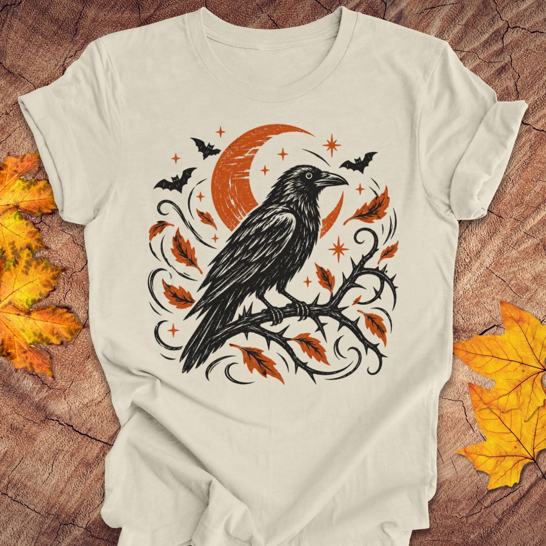 An off-white t-shirt featuring a Halloween graphic of a black raven on a branch with orange leaves and a crescent moon, laid flat on a rustic wood background with autumn leaves.