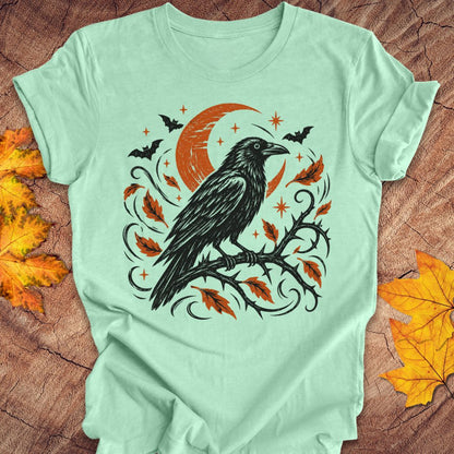 A mint green short-sleeve t-shirt with a Halloween graphic of a black raven on a branch with orange leaves and a crescent moon, laid flat on a rustic wood background with autumn leaves.