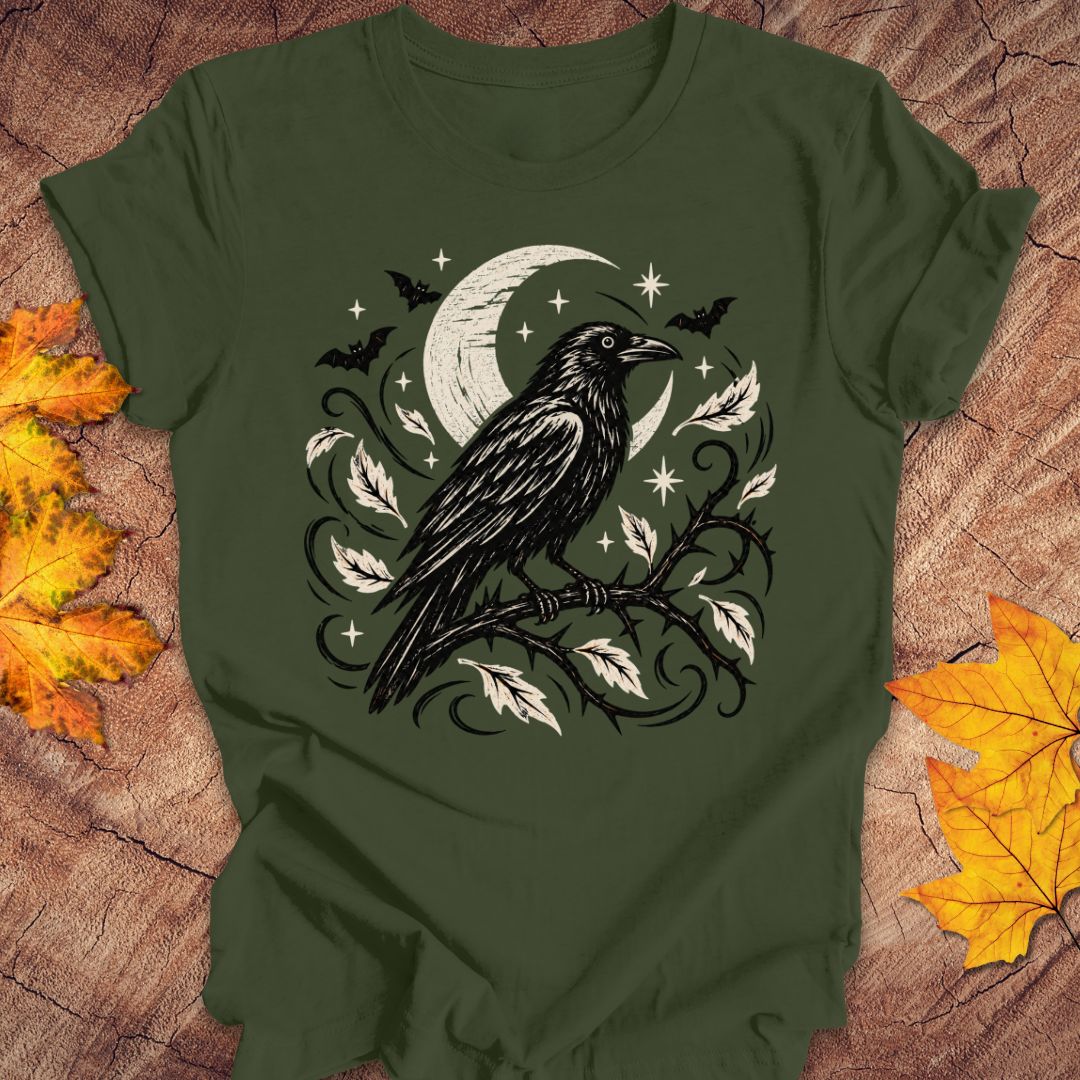 An olive green short-sleeve t-shirt with a black and white graphic of a raven perched on a branch under a crescent moon and stars, styled flat on a wood background with autumn leaves.