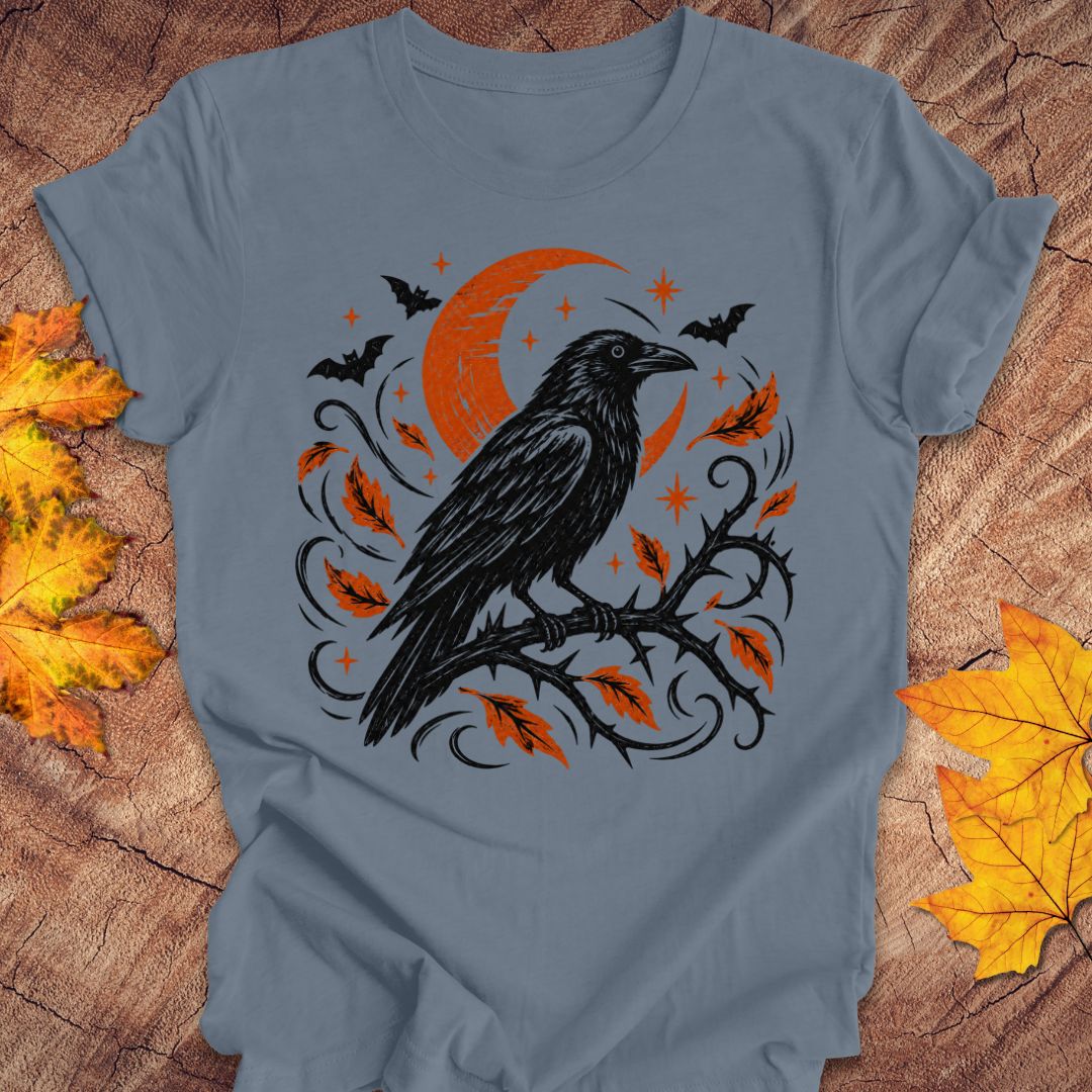A slate blue t-shirt featuring a Halloween graphic of a black raven on a branch with orange leaves, a crescent moon, and bats, laid flat on a rustic wood background with autumn leaves.