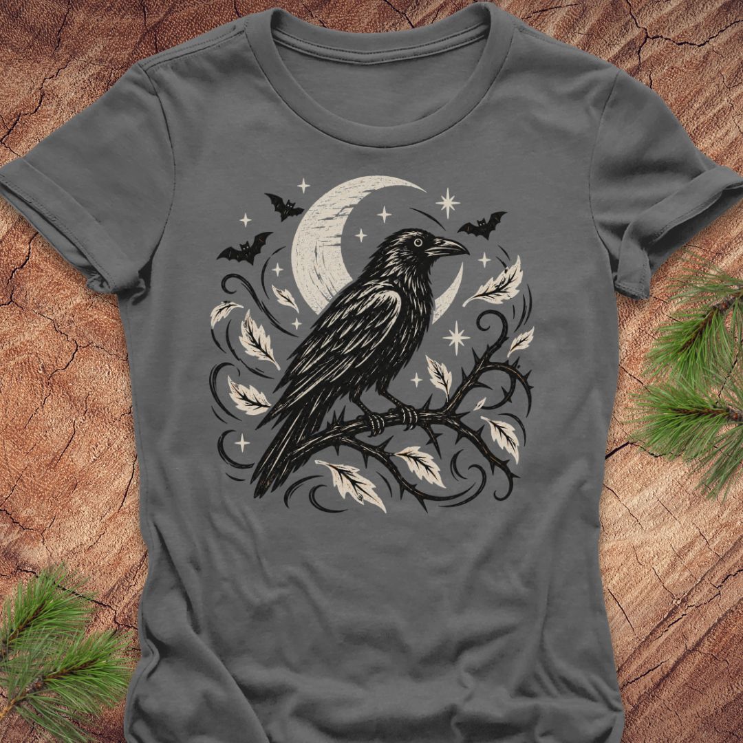 A dark gray short-sleeve t-shirt with a detailed graphic of a black raven perched on a branch with white leaves, set against a crescent moon and stars. The shirt is laid flat on a rustic wood background with pine needles in the corners.