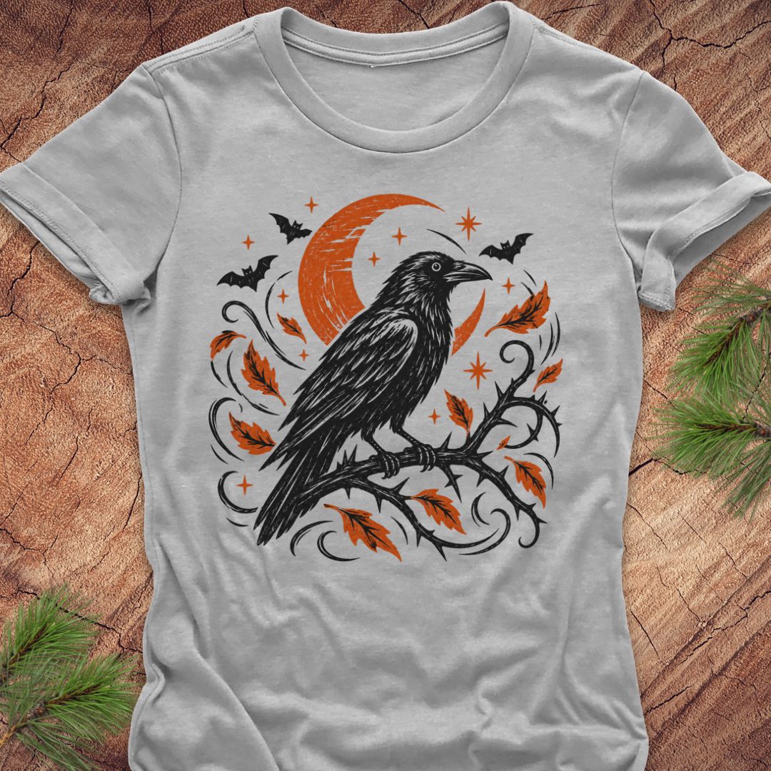 A light gray short-sleeve t-shirt with a Halloween graphic print of a black raven on a branch with orange leaves, a crescent moon, and bats, laid flat on a rustic wood background with pine needles.