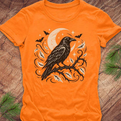An orange short-sleeve crewneck t-shirt featuring a black and cream graphic of a raven on a branch with a crescent moon, stars, and bats, laid flat on a rustic wood background with pine needles.