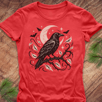 A red short-sleeve crewneck t-shirt featuring a black and white graphic of a raven perched on a branch with a crescent moon and stars, laid flat on a rustic wood background with pine needles.