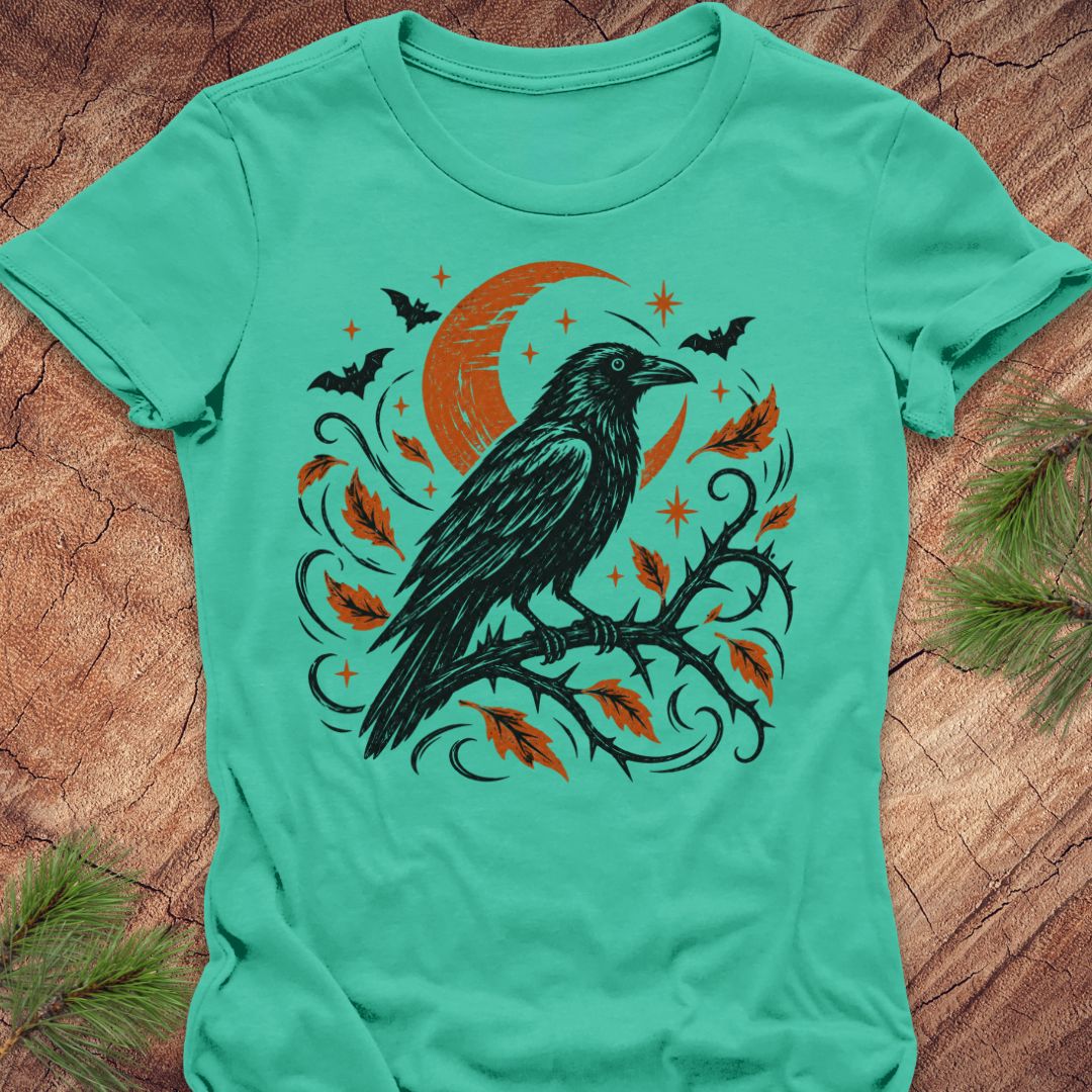 A mint green short-sleeve t-shirt with a Halloween graphic of a black raven on a branch with orange leaves and a crescent moon, laid flat on a rustic wood background with pine needles.