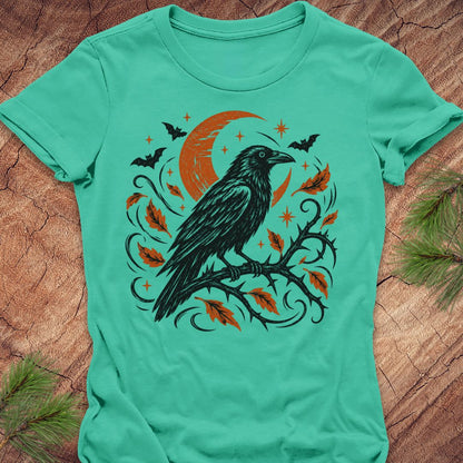 A mint green short-sleeve t-shirt with a Halloween graphic of a black raven on a branch with orange leaves and a crescent moon, laid flat on a rustic wood background with pine needles.