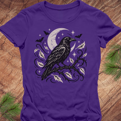 A purple short-sleeve crewneck t-shirt featuring a gothic graphic of a black raven perched on a branch with a crescent moon, stars, and leaves, laid flat on a rustic wood background with pine needles.