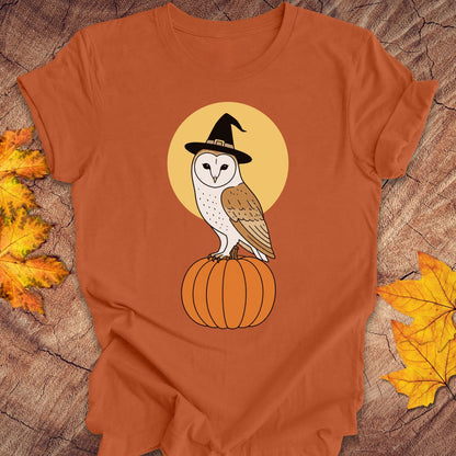 An orange short-sleeve t-shirt laid flat on a rustic wood surface, featuring a graphic of a white barn owl wearing a black witch hat perched on an orange pumpkin in front of a yellow circle. Yellow and orange autumn leaves are placed on either side of the shirt.