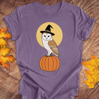 A purple short-sleeve t-shirt laid flat on a rustic wood surface with autumn leaves. The shirt features a graphic of a white and brown barn owl wearing a black witch hat perched on an orange pumpkin, set against a yellow circle.