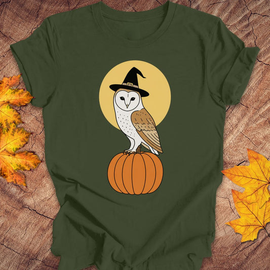 An olive green short-sleeve t-shirt with a graphic of a barn owl in a witch hat perched on a pumpkin against a yellow circle, styled flat on a wood background with autumn leaves.