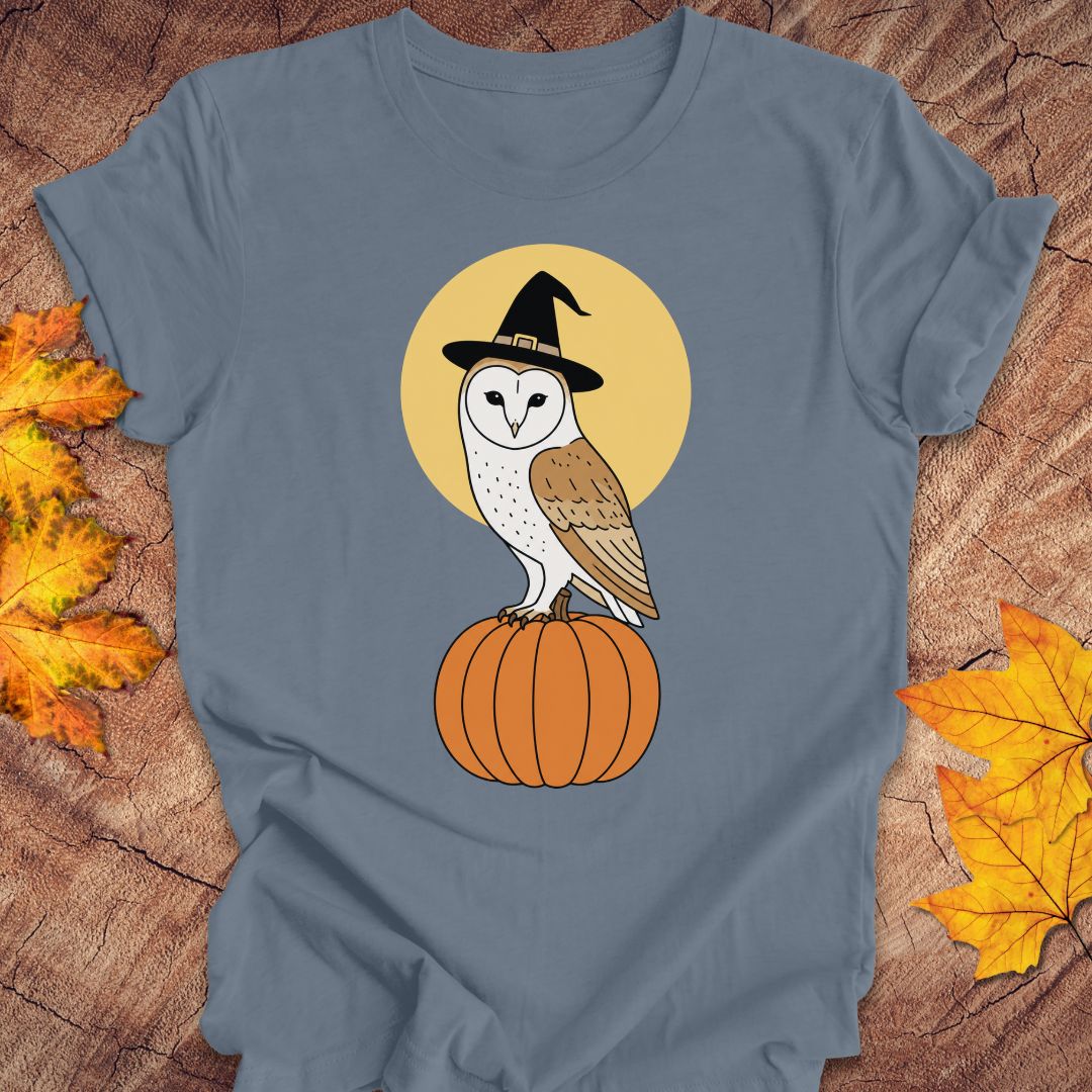 A slate blue t-shirt with a graphic of a barn owl in a witch hat perched on a pumpkin against a yellow moon, styled on a wood background with autumn leaves.