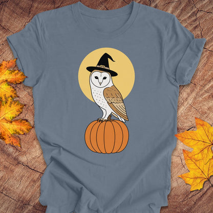 A slate blue t-shirt with a graphic of a barn owl in a witch hat perched on a pumpkin against a yellow moon, styled on a wood background with autumn leaves.