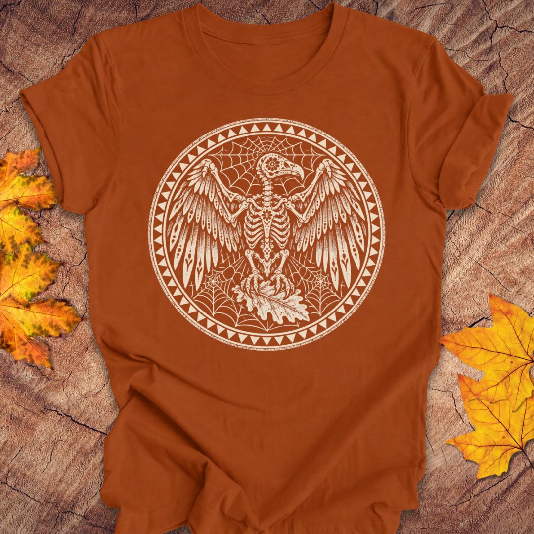 An orange-brown short-sleeve t-shirt with a circular graphic of a skeletal bird surrounded by autumn leaves and webs, laid flat on a wood surface with yellow and orange leaves around it.