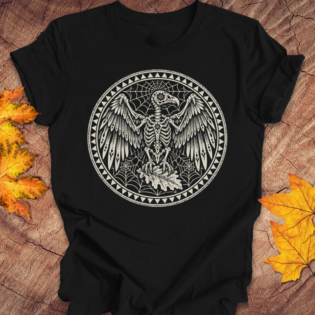 A black short-sleeve t-shirt with a circular graphic of a skeletal bird surrounded by webs, laid flat on a wood surface with autumn leaves.